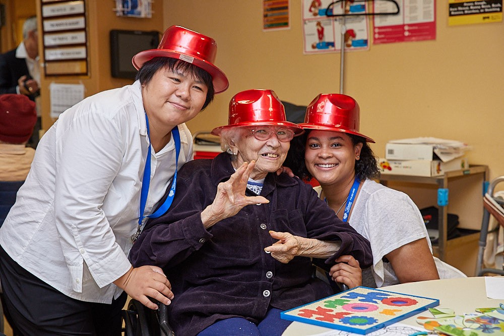 Three smiling individuals, including an elderly woman in a wheelchair, wearing festive red hats, celebrate holidays together in a cheerful indoor setting with decorations and crafts on the table