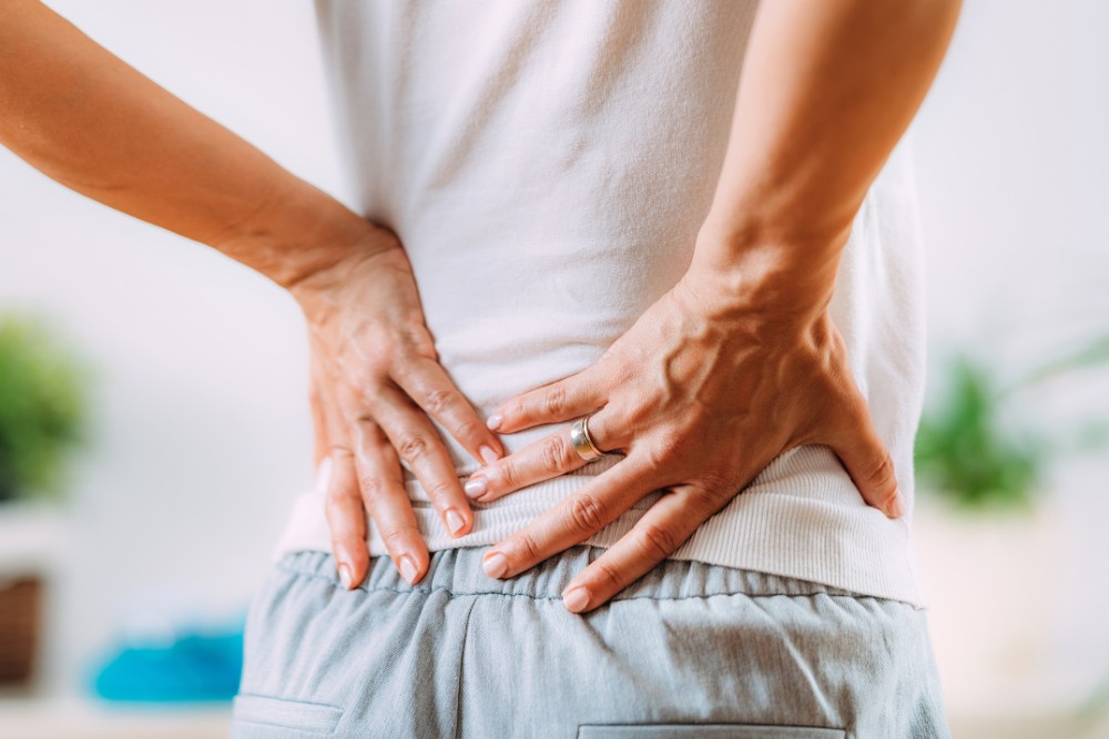 Person experiencing chronic sciatica pain, holding their lower back and upper leg in discomfort, dressed in casual clothing, indoors.