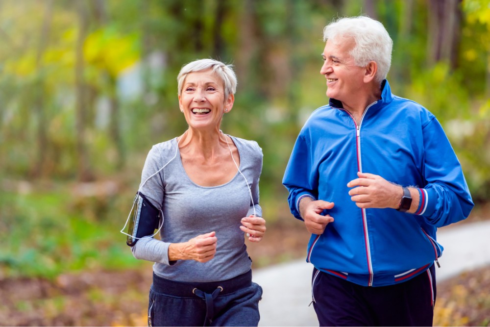 Active senior couple jogging outdoors in a park, smiling and enjoying exercise. A reminder of the benefits of staying active and avoiding the negative effects of a lack of exercise.